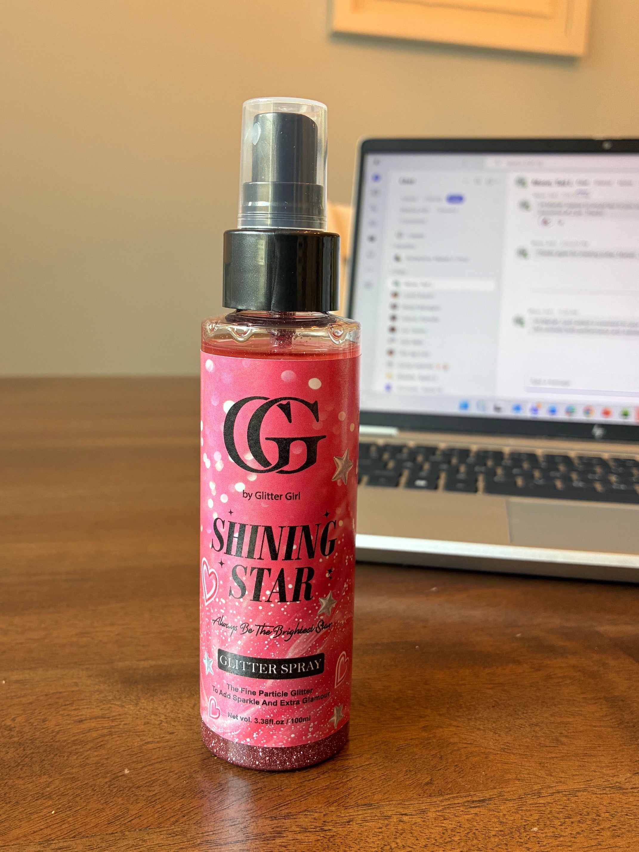 A bottle of glitter spray by Glitter Girl, labeled 'Shining Star', stands on a wooden table with a blurred laptop in the background.
