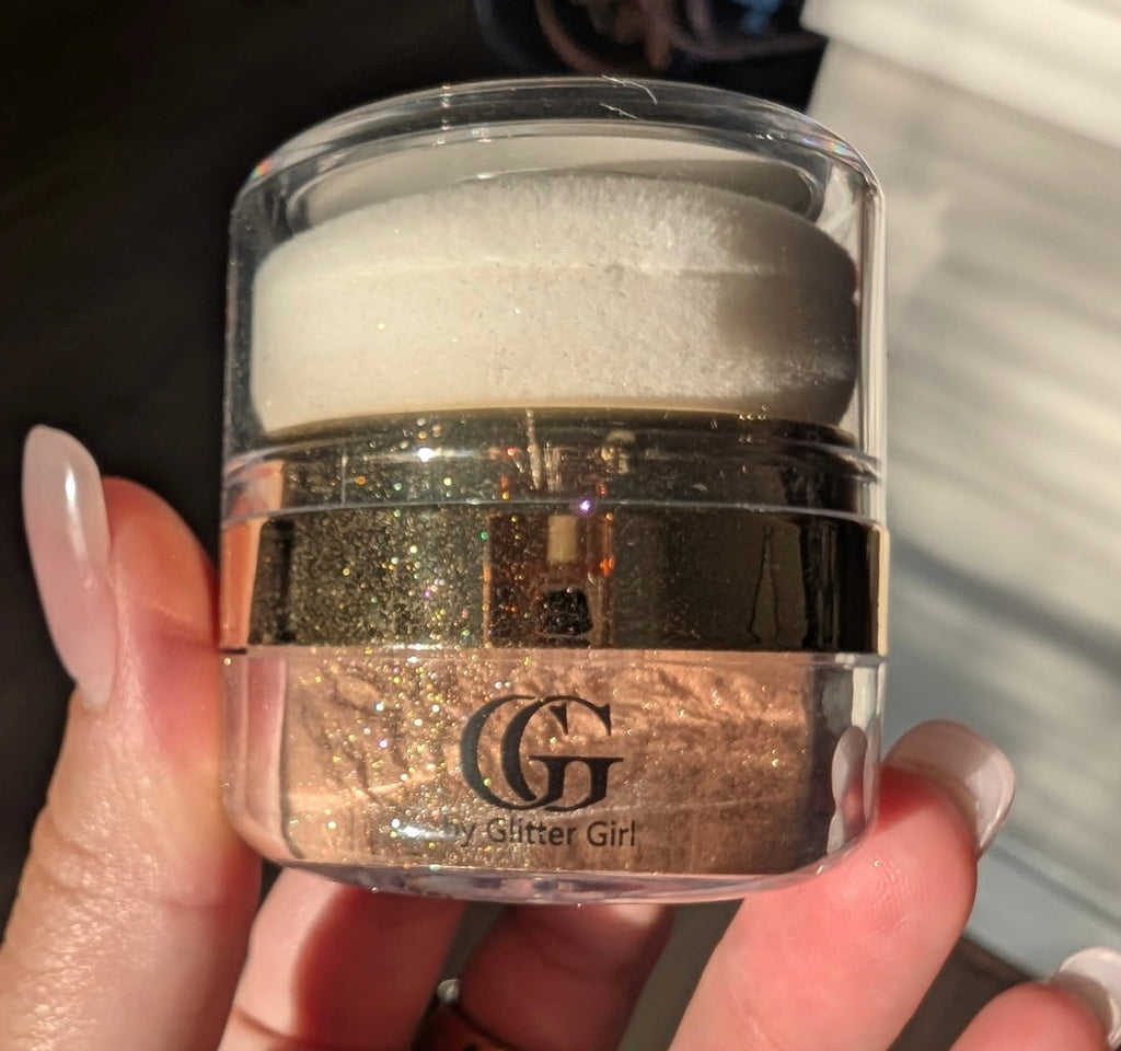 A hand holding a small clear cosmetic jar filled with shimmering rose-gold glitter powder, with a white sponge on top and the GG My Glitter Girl logo printed on the front.